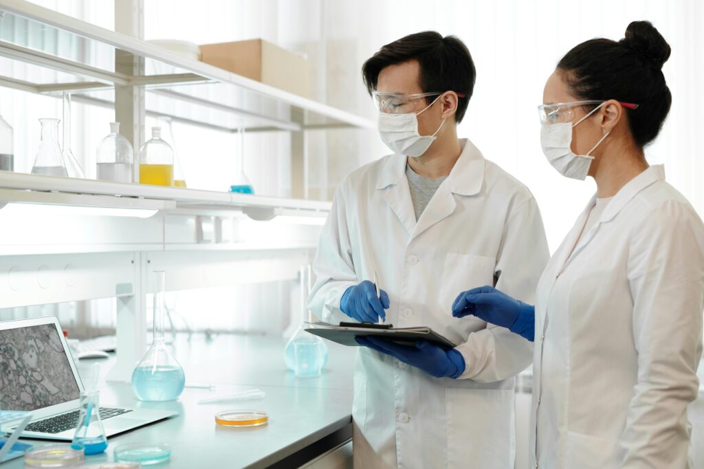 A male and female researchers in PPE look at results and findings in a laptop in a lab setting with labware and equipment around them.